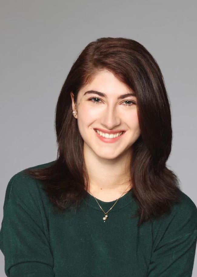 Bridget Disini, with her long dark hair cascading over a green sweater, smiles warmly at the camera. Her necklace catches the light, adding a touch of elegance against the subtle gray background.