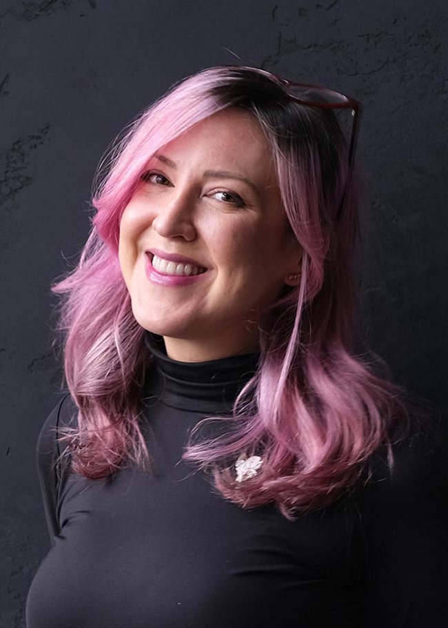 Dr. Cara Dunkley, sporting vibrant pink hair, smiles warmly while wearing a black turtleneck against a dark background.