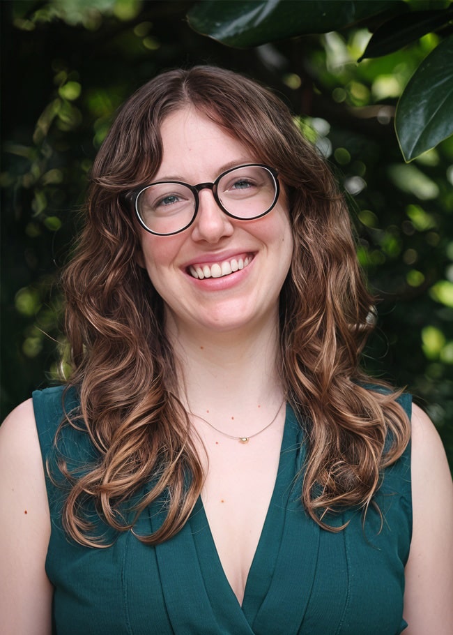 Dr. Eva Harris, a renowned research scientist with long curly hair, wears glasses and a green sleeveless top as she smiles at the camera. The backdrop of lush greenery complements her vibrant presence.