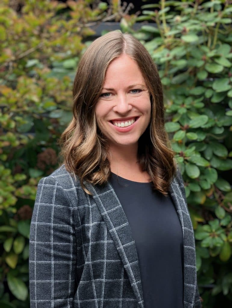 Dr. Heather Morton, a woman with long brown hair, wearing a gray checkered blazer and black top, stands smiling in front of green foliage.
