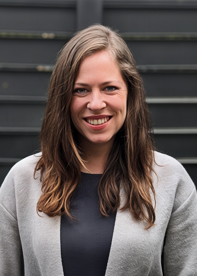 Dr. Heather Morton, with long hair flowing, smiles warmly while standing in front of a dark fence, wearing a stylish gray jacket.