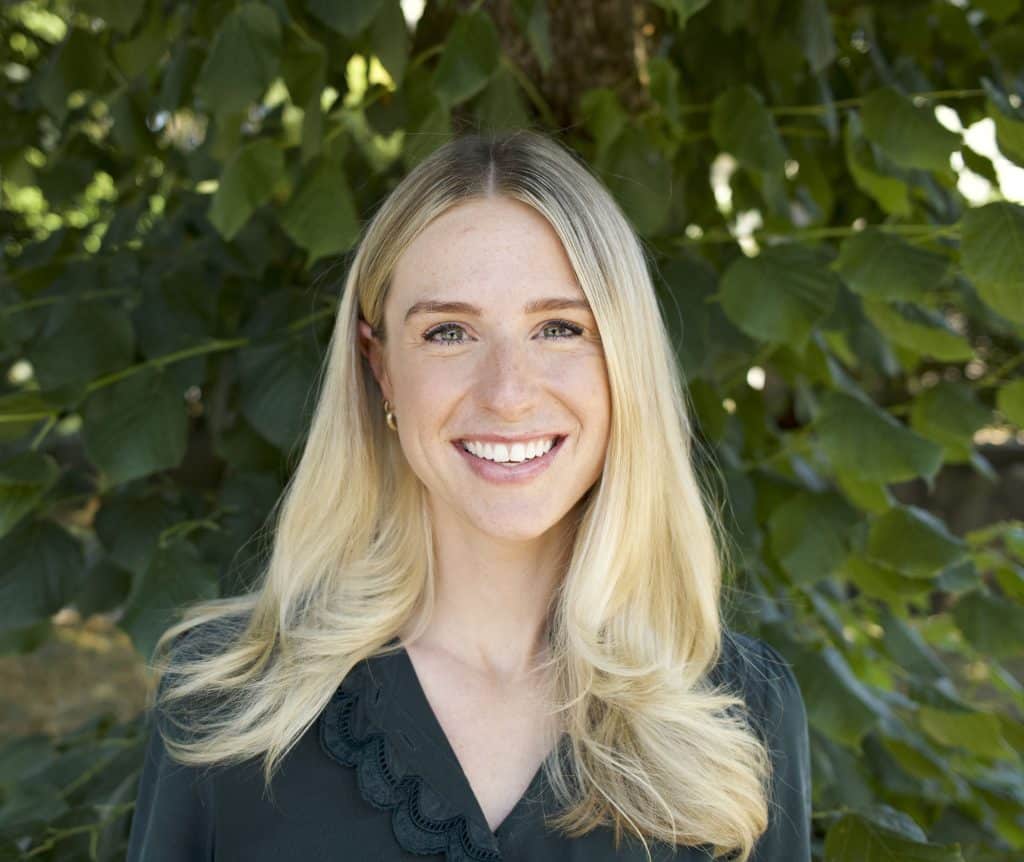Dr. Julia Poole, a woman with long blonde hair, smiles in front of a leafy green tree, wearing a dark blouse.