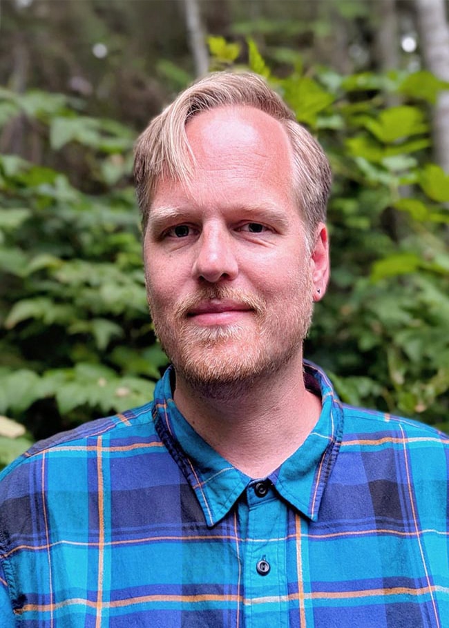 Dr. Jan van der Tempel, a person with a beard and short, light hair, stands outdoors wearing a blue plaid shirt, surrounded by the lush backdrop of trees.