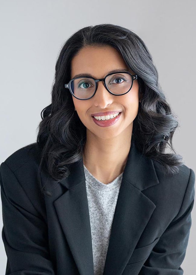 Dr. Janani Sankar, with long, dark hair and glasses, sits smiling in a black blazer and gray shirt, her hands clasped elegantly.