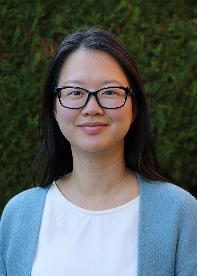 Dr. Jane Woo, with her glasses and long hair, stands gracefully in front of a green, leafy background, wearing a white shirt and blue cardigan.