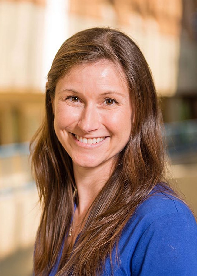 Dr. Joelle LeMoult, with her long hair flowing gracefully, smiles into the camera while standing indoors against a beautifully blurred background.