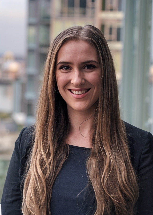 Dr. Katerina Rnic, with her long brown hair cascading over a black jacket, stands on a balcony overlooking the city buildings.