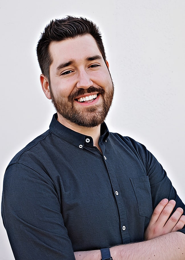 Mark Antczak, a man with a beard and short hair, stands smiling with arms crossed. He's clad in a dark button-up shirt and sports a watch. The plain white background highlights his confident demeanor.