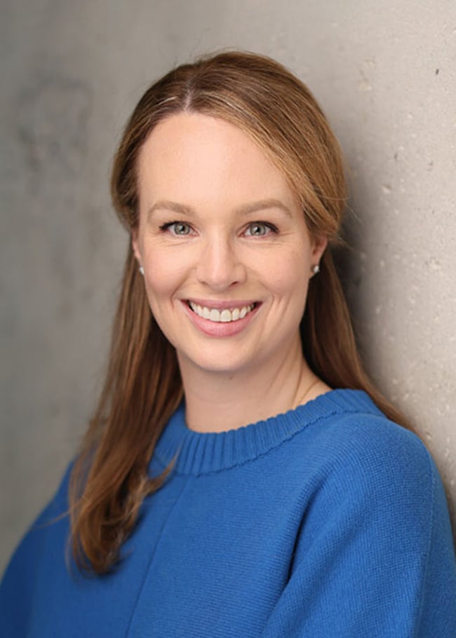 Dr. Elizabeth Peters, with long hair and a blue sweater, smiles while leaning against a textured wall.