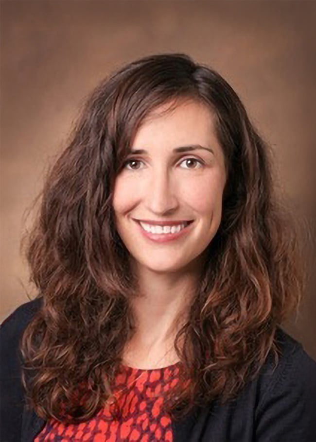 Dr. Sonia Matwin, with her long, wavy brown hair, radiates warmth in a red patterned top and black cardigan, smiling against a neutral background.