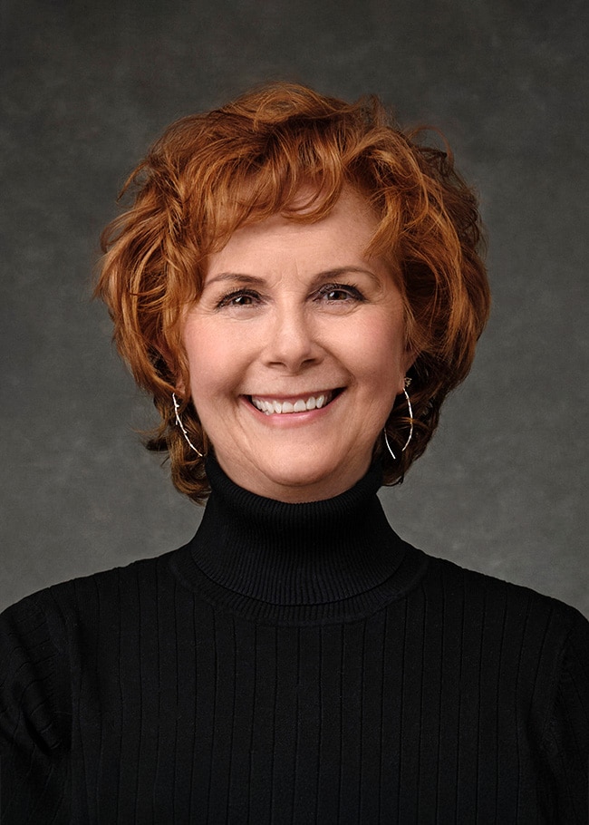 Dr. Maureen Whittal, with her curly red hair, smiles warmly in a black turtleneck against a gray background.