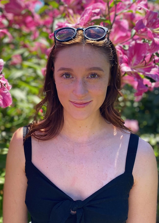 Brett stands with sunglasses on her head in front of vibrant pink flowers, wearing a black top, outdoors.