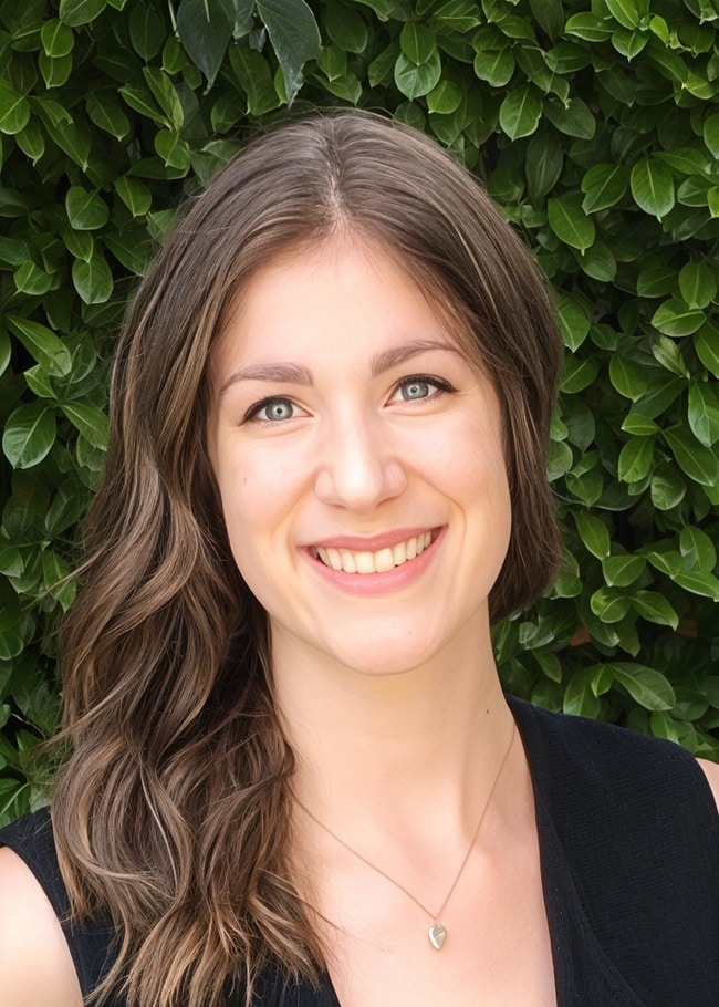 A person with long brown hair smiles in front of a leafy green background.