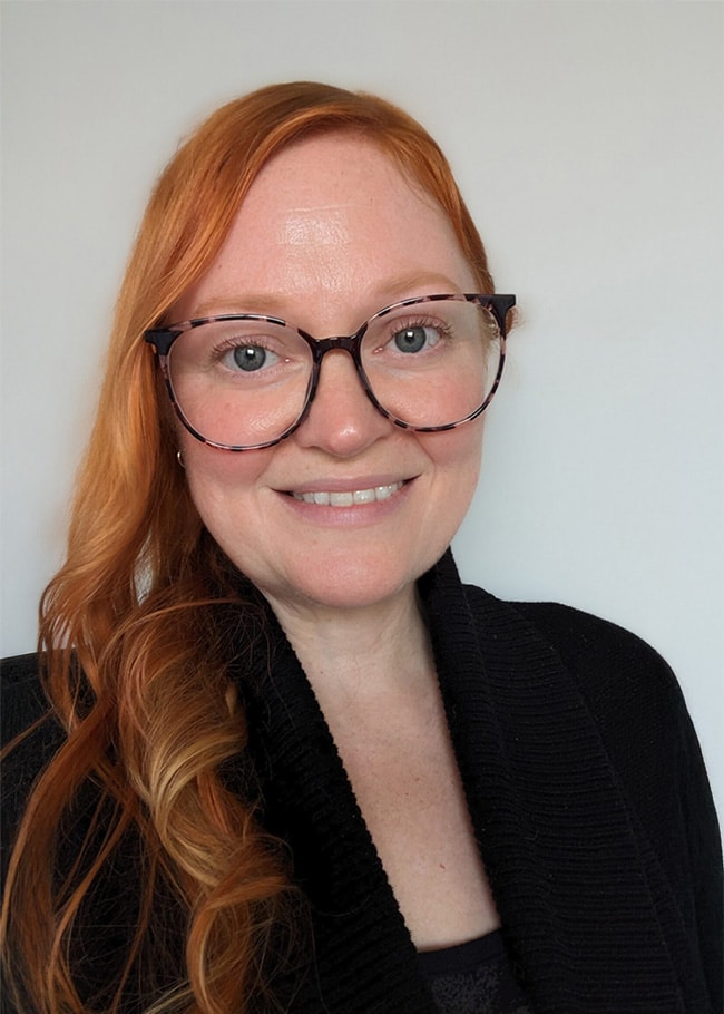 A person with red hair and glasses smiles at the camera, wearing a black top against a plain background.