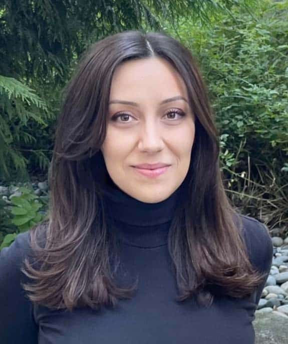 Mathilde Rioux, a woman with long dark hair wearing a black turtleneck, stands outdoors in front of green foliage and rocks, facing the camera and smiling slightly.