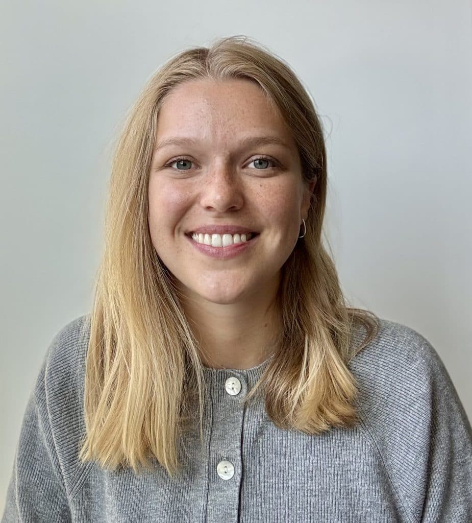 Mathilde Rioux, with straight blonde hair, wearing a grey sweater and small hoop earrings, smiles at the camera against a plain light background.