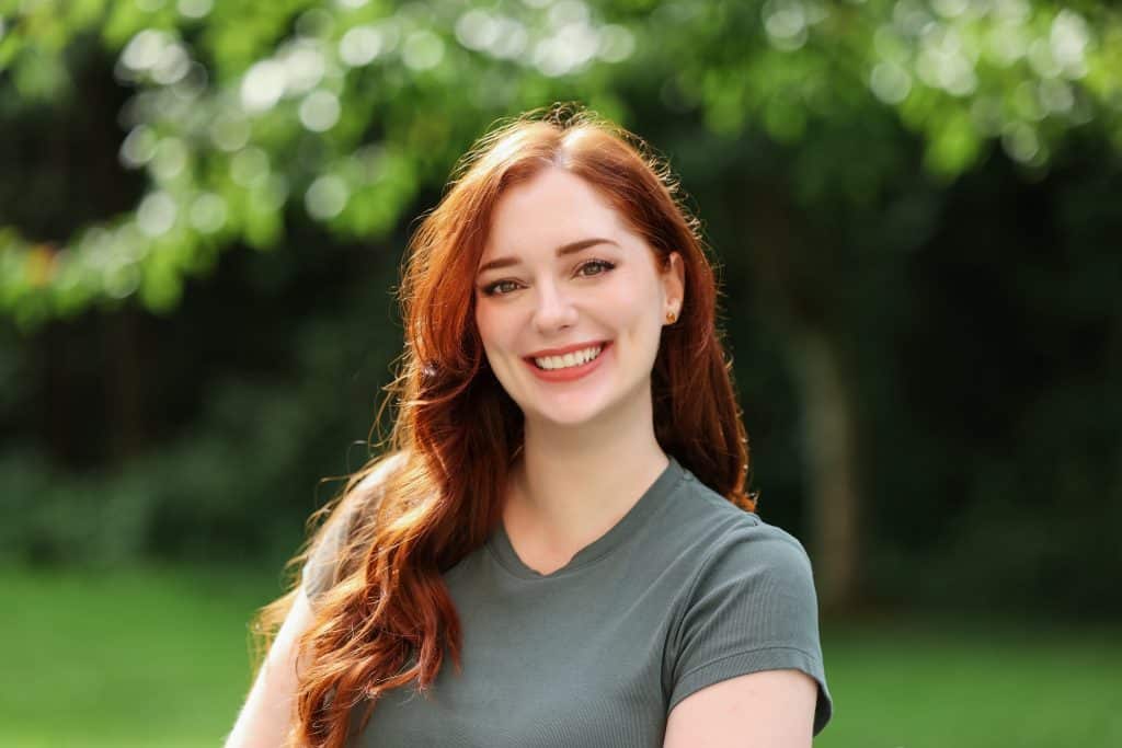 A woman with long red hair wearing a gray t-shirt smiles outdoors with green trees blurred in the background.