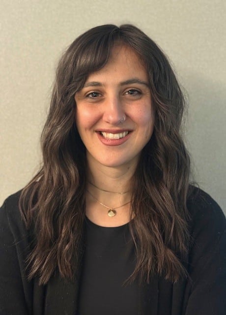 Dr. Siobhan Fitzpatrick, a woman with wavy brown hair, wearing a black top, black cardigan, two necklaces, and a nose ring, smiles at the camera against a plain light background.