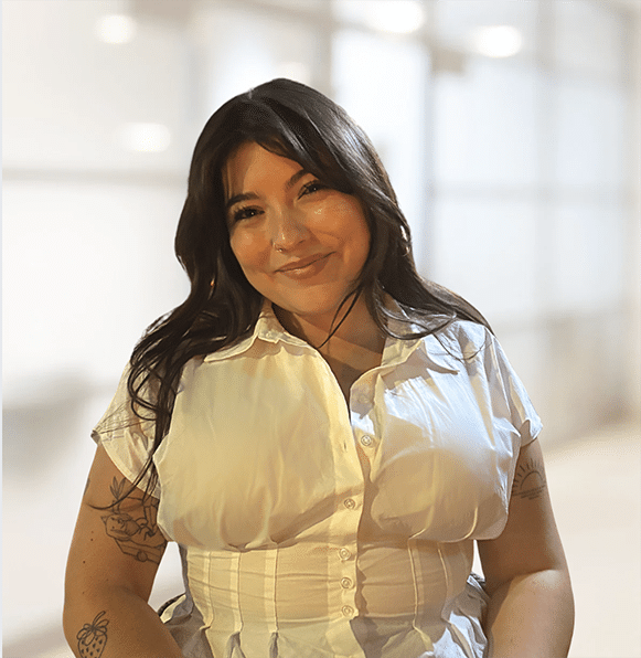 Erin Fitzpatrick, a woman with long dark hair and tattoos on both arms, smiles at the camera in a short-sleeved white blouse. The background is softly blurred.