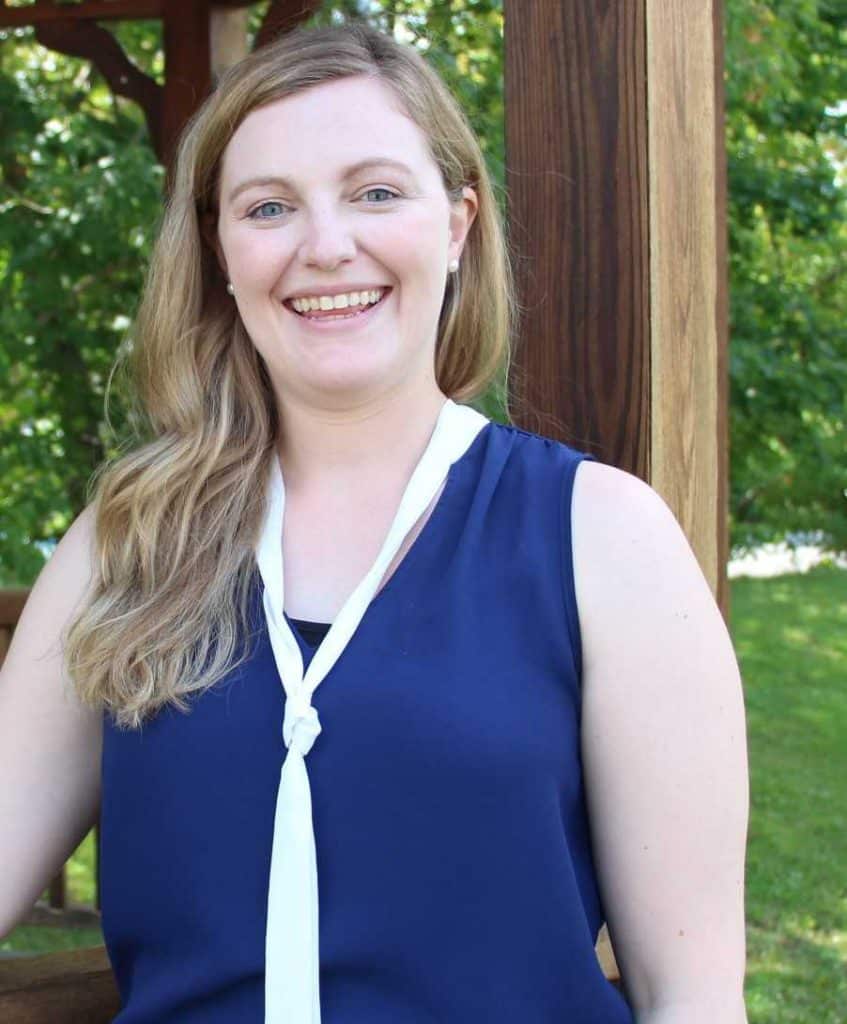 Dr. Kate Kysow, a woman with long blonde hair, wearing a navy sleeveless top and white scarf, stands outdoors smiling in front of wooden posts and green foliage.