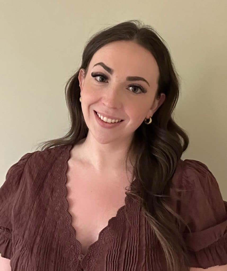 Woman with long brown hair, wearing a mauve blouse, stands in front of a plain light-colored wall and smiles at the camera.